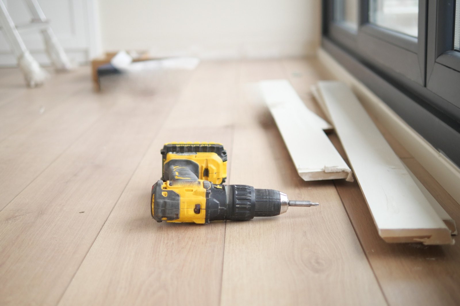 Yellow and black cordless drill rests on wooden flooring beside scattered materials.