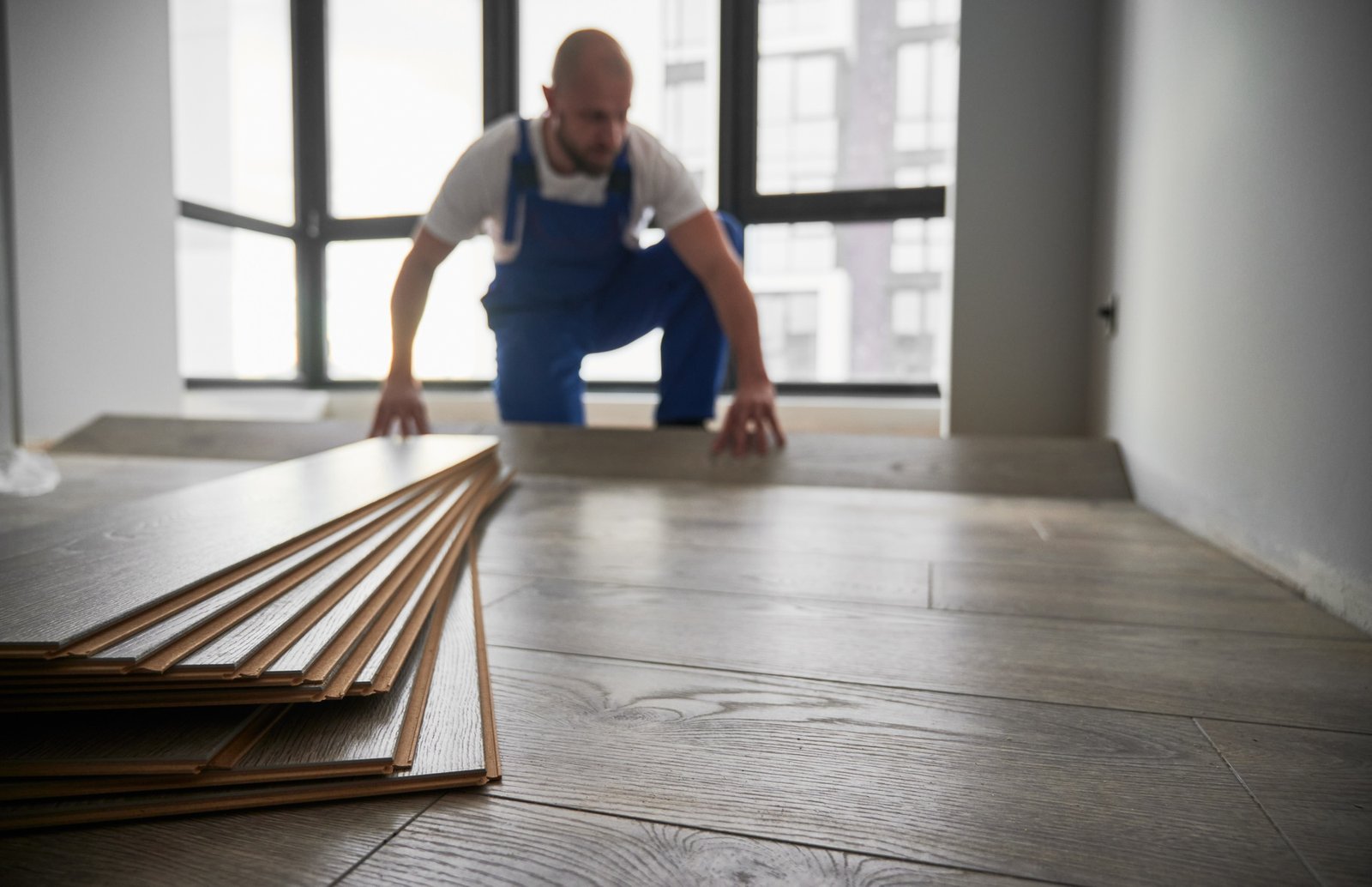 A worker in blue overalls lays wooden flooring in a bright, modern room.