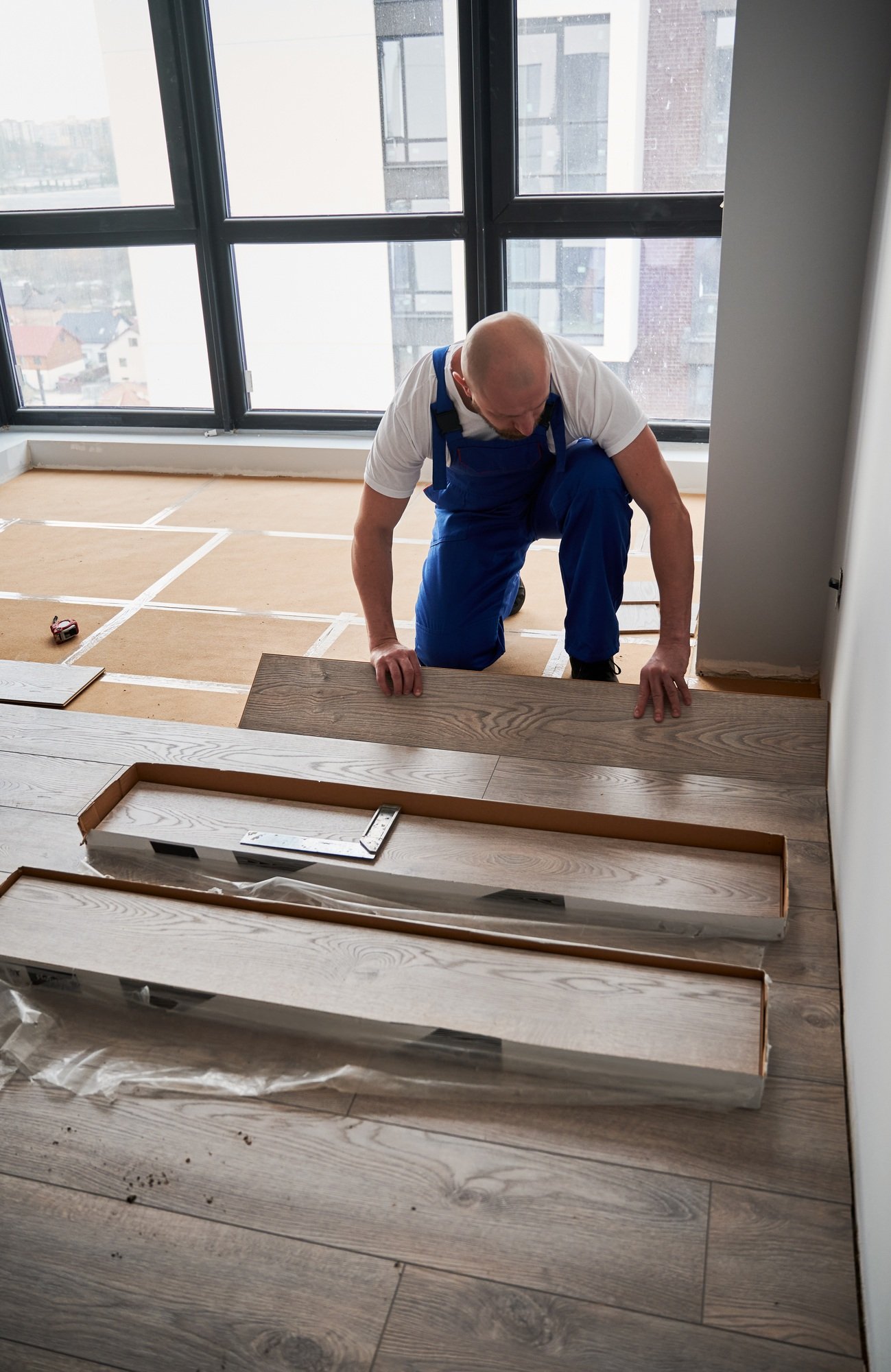A worker installs wood-like flooring in a bright room with large windows.