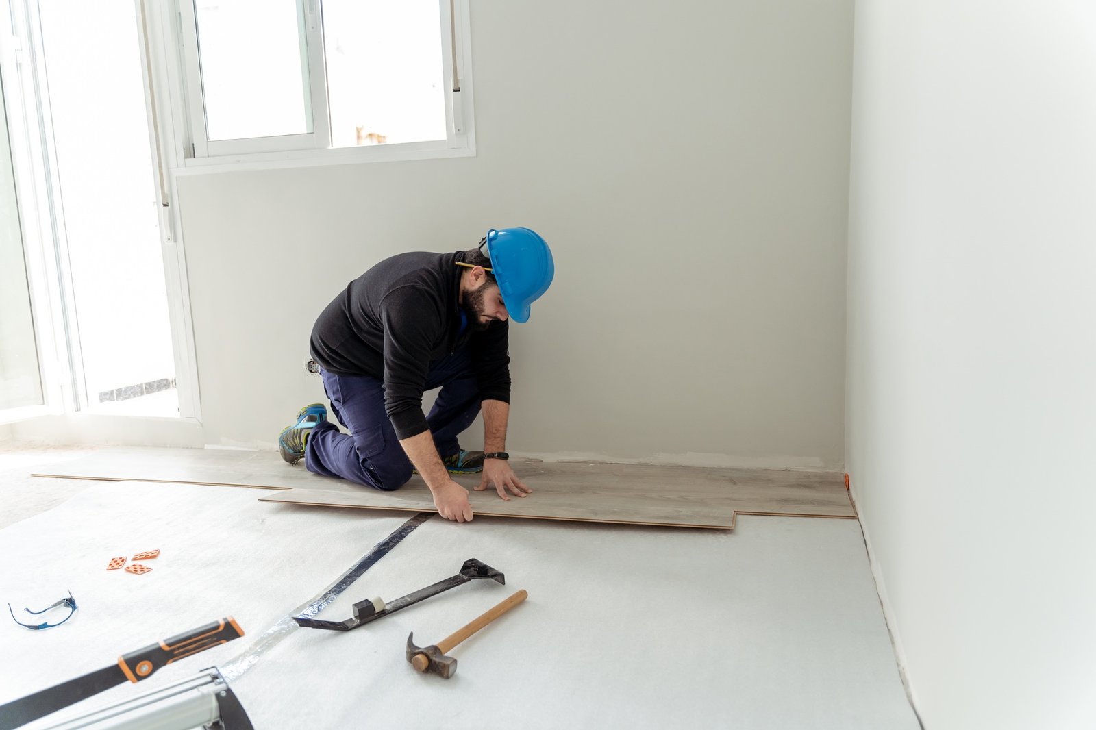 Man wearing a blue hard hat installs wooden flooring in a bright, empty room.