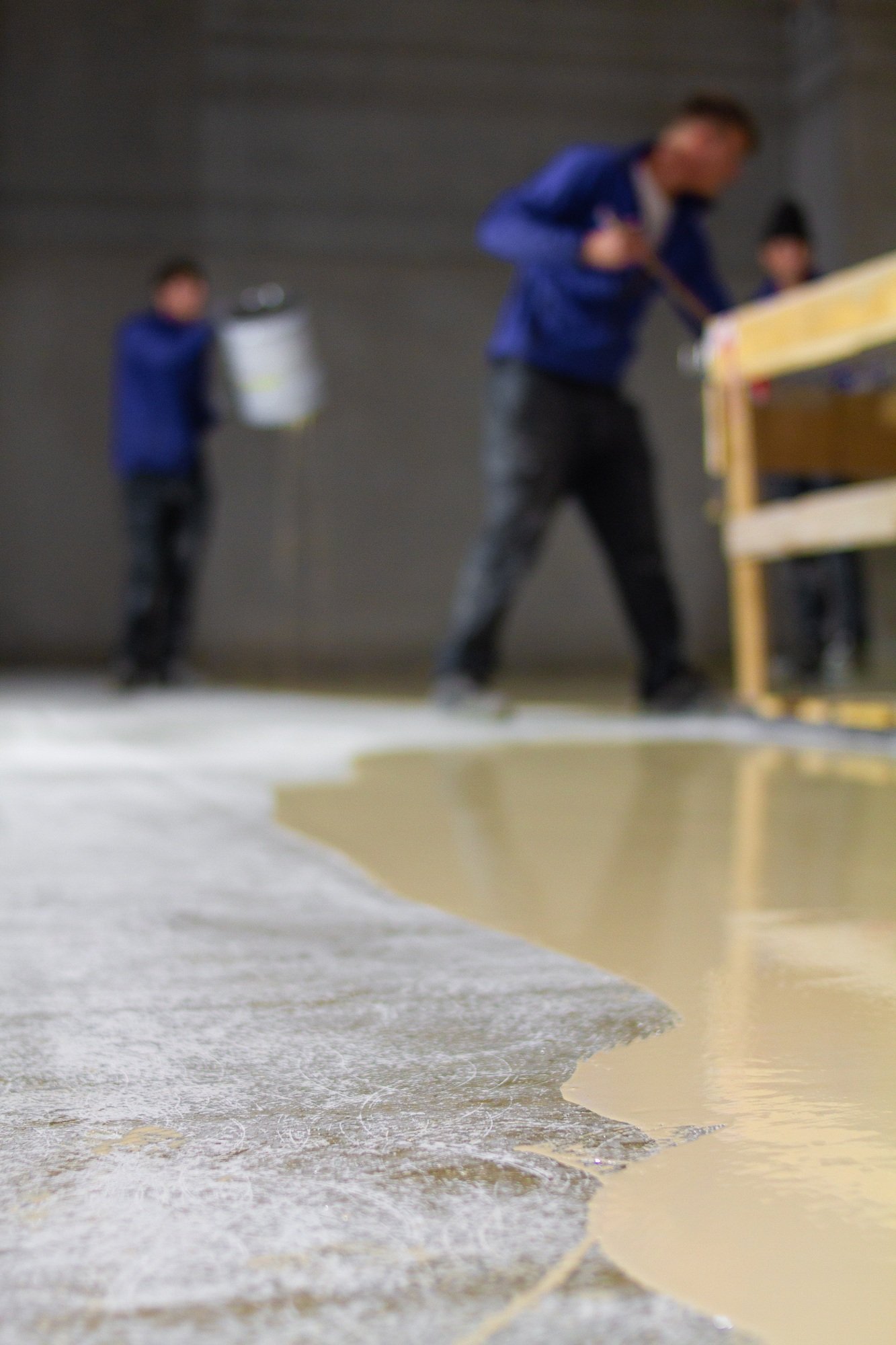 Workers apply a smooth, light-colored liquid to a concrete floor in a construction setting.