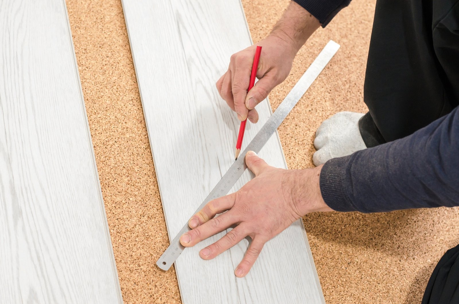 Person measuring and marking a white wooden plank on a cork surface with a ruler and pencil.